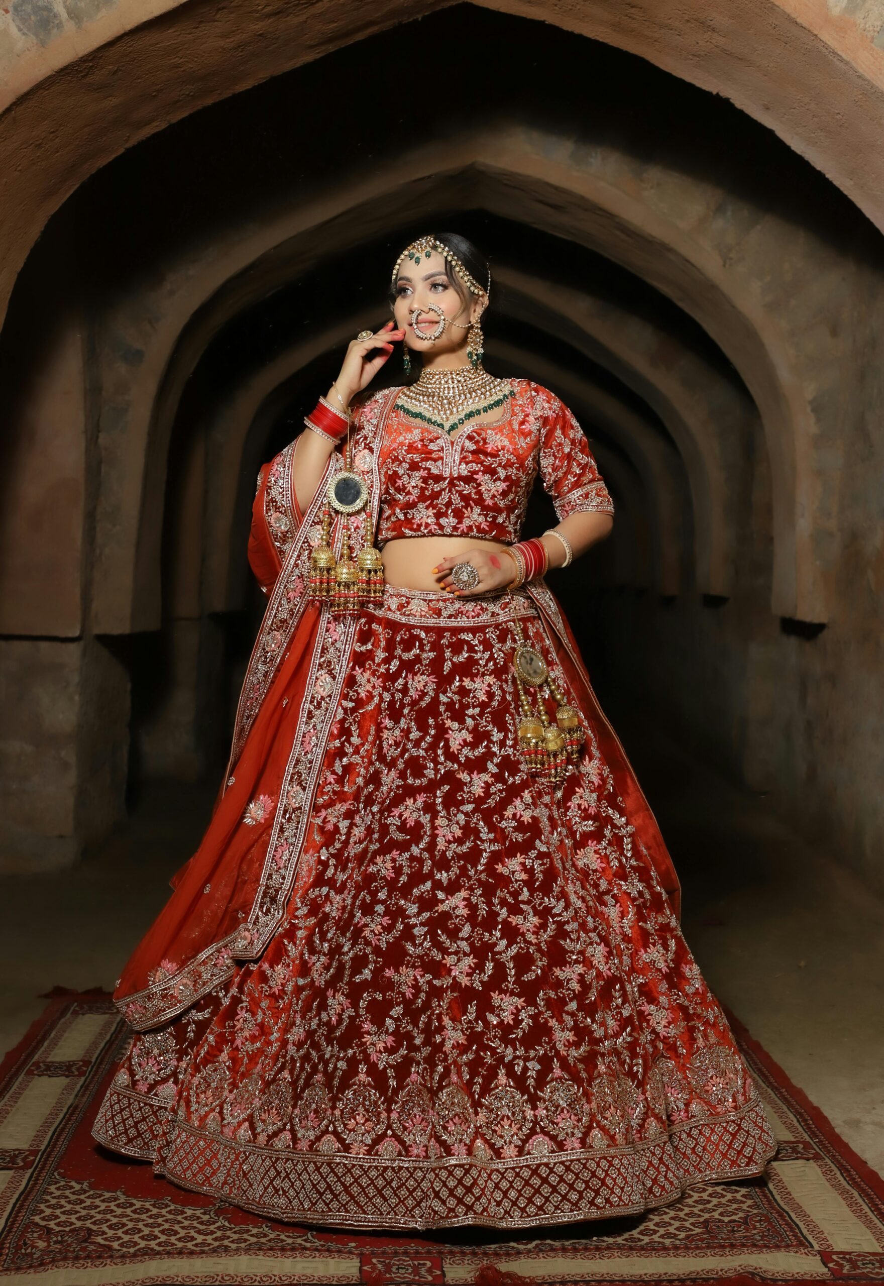 A beautiful Indian bride in a red lehenga, posing gracefully indoors with intricate jewelry.