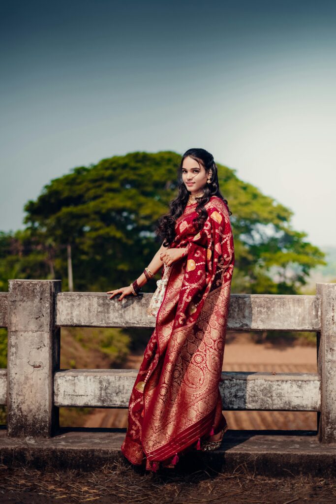 A woman wearing a red ornate saree stands outdoors by a fence, embracing cultural elegance.