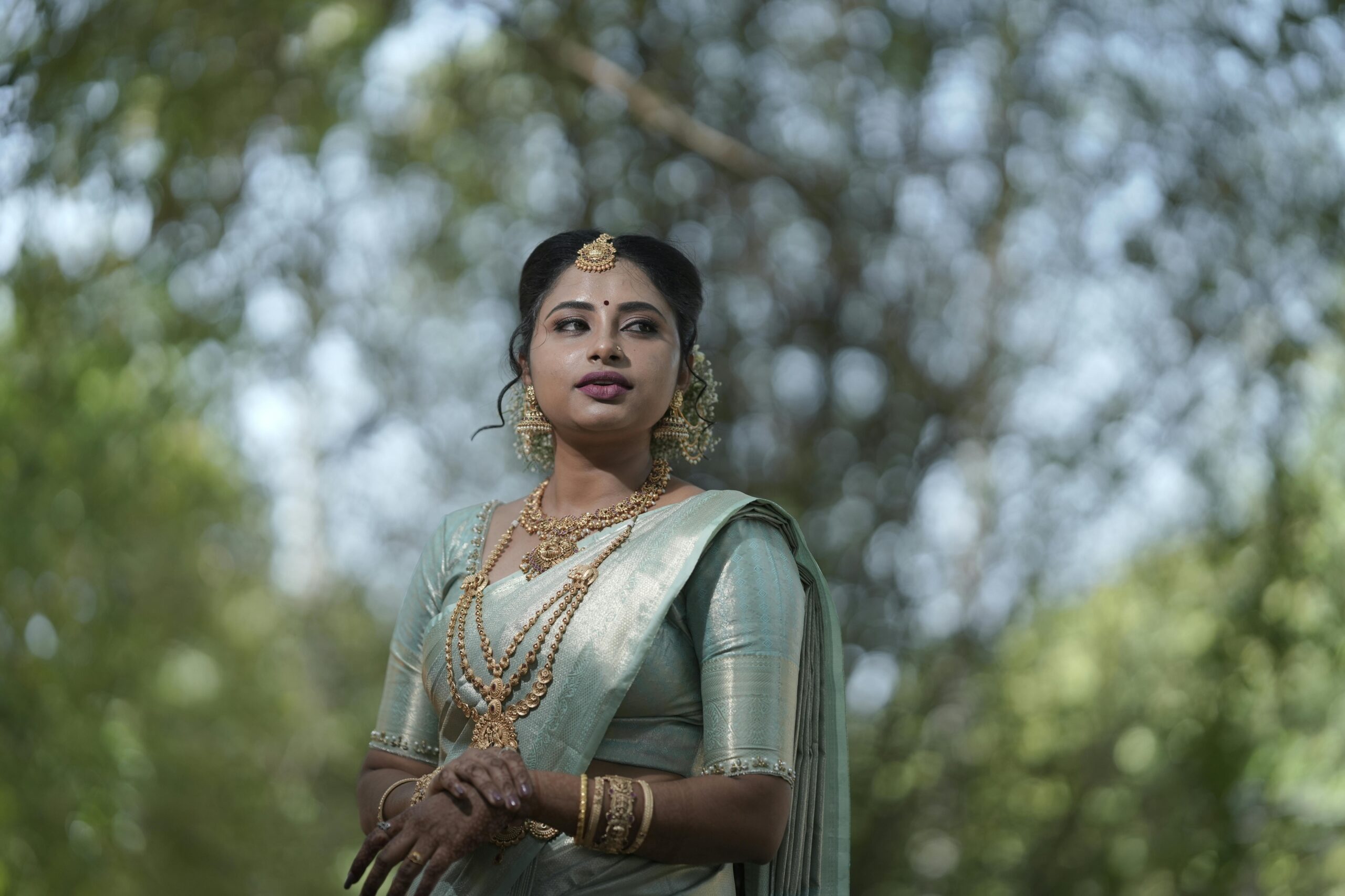 A beautiful woman in a pista green saree poses elegantly in an outdoor setting with lush greenery.