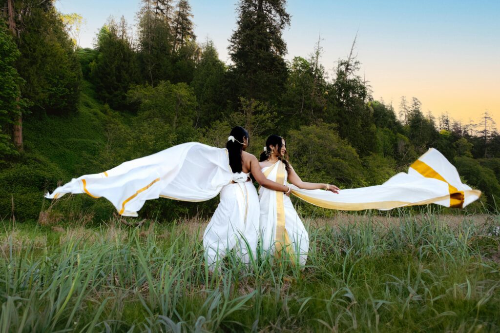Two women in white sarees with flowing fabric in a lush, outdoor setting under a colorful sky.
