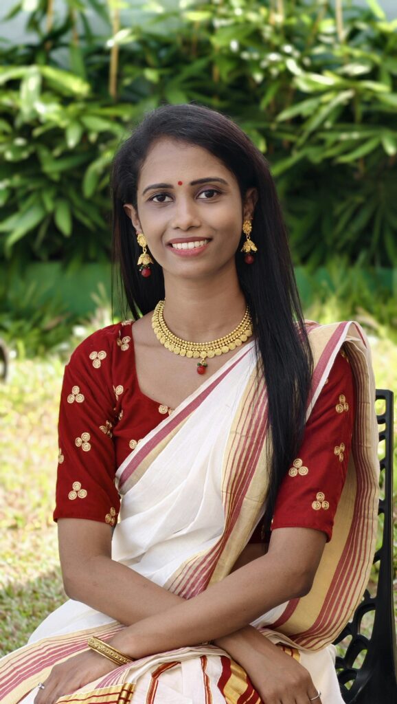 Smiling South Asian woman in traditional Kerala saree sitting outdoors, showcasing vibrant cultural attire.