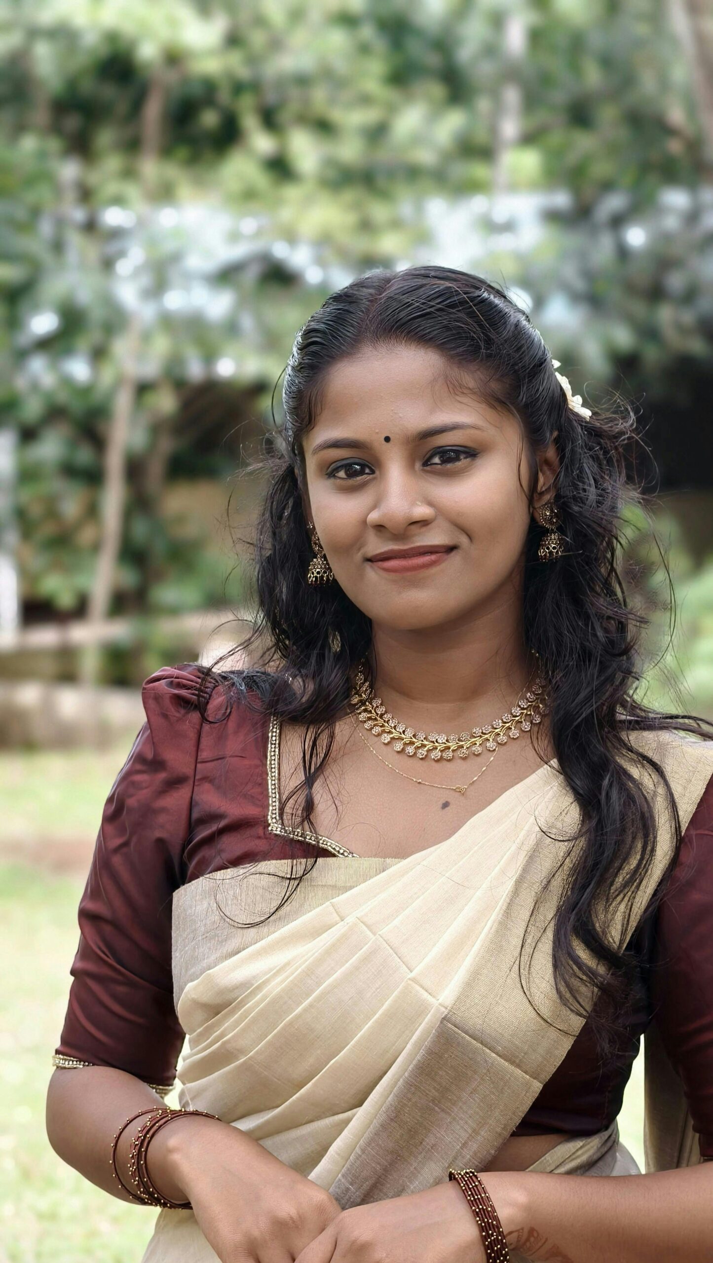 A young woman in a Kerala saree, smiling outdoors, wearing traditional jewelry.